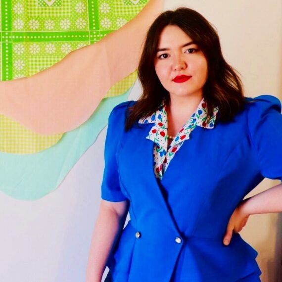 Photo of a white woman in a blue blazer. She has brown hair and is standing in front of artwork.