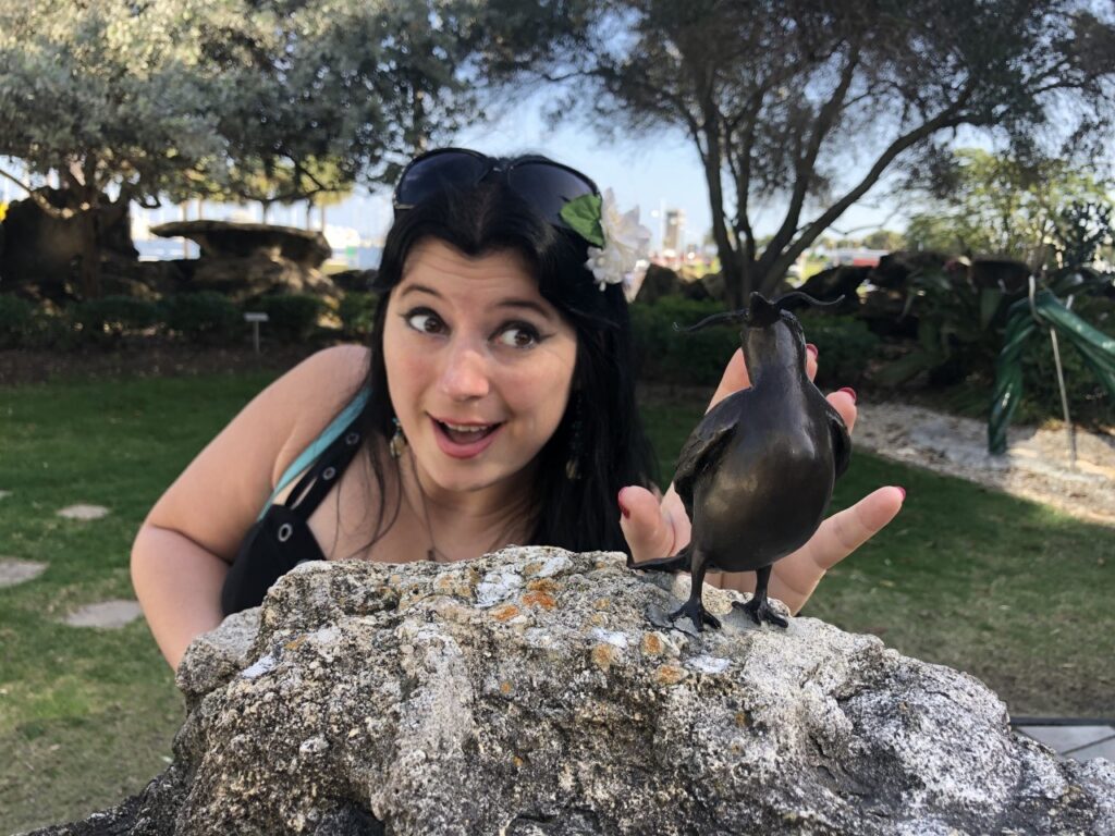 Erica Pastecki pictured petting the miniature statue of a black bird perched on a granite rock. Erica is caucasion, pale with freckels. She is wearing a black dress and dark sunglasses. she has long jet black hair.