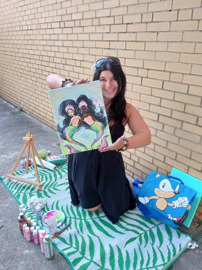 Erica Pastecki sitting on a stripped blanket outside in front of a tan bricked wall. She is surrounded by artwork, paints and an easel. She is holding up a completed painting of a pair of mermaids. Erica is white, pale with freckles, wearing a black dress and dark sunglasses. She is smiling.