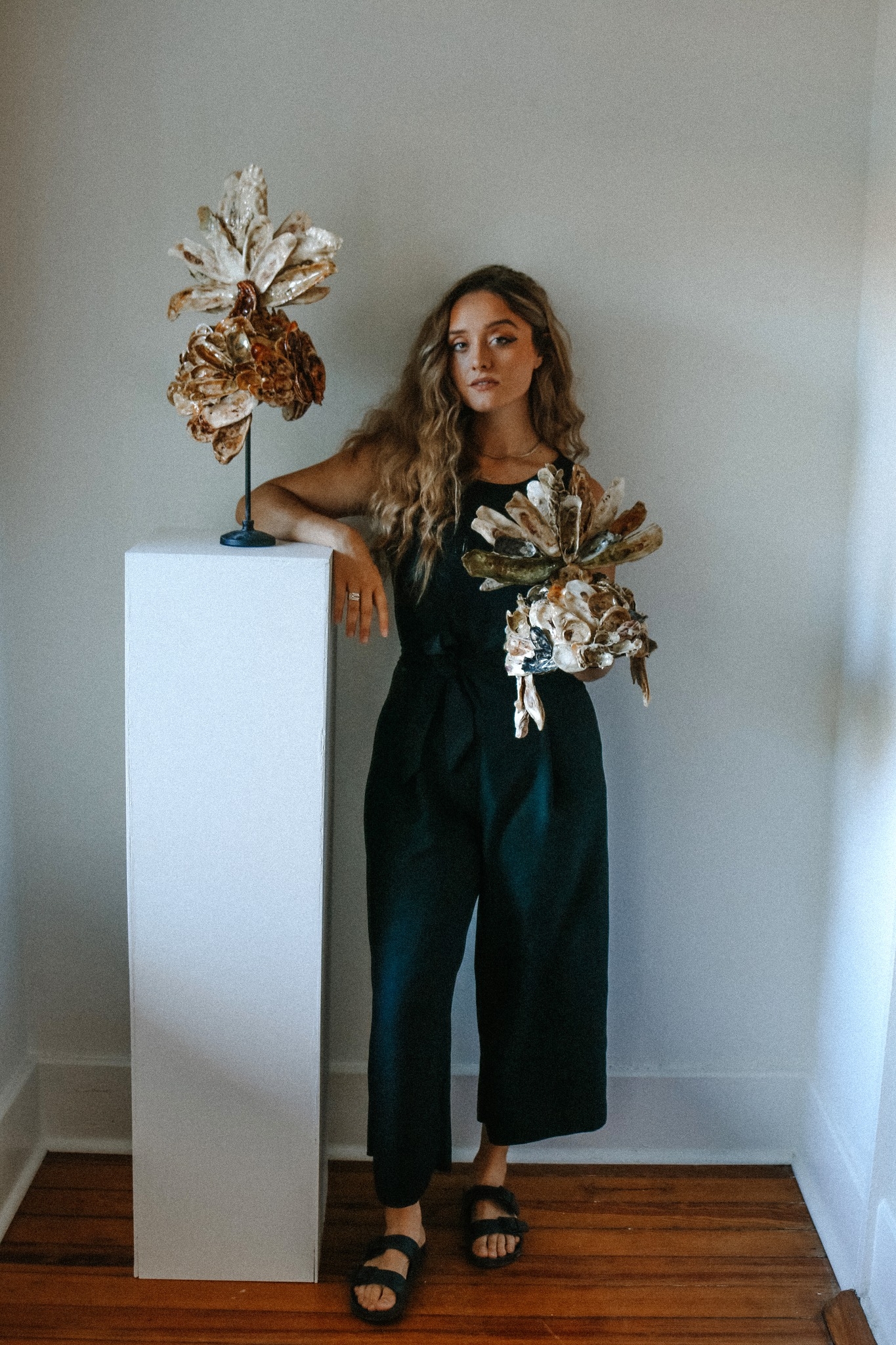 Stephanie Forbes standing next to her work "Divine Armour and the Lowcountry". Stephanie is wearing a black jumpsuit and sandals, she has long curly blonde hair and pale sin. She is standing with her two romanesque battle helmets, which are made of recycled oyster shells. All infront of a white wall.