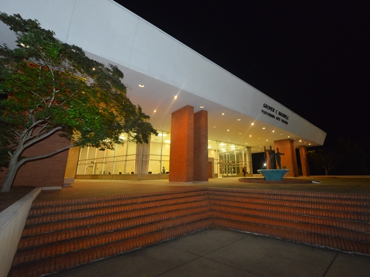 photo of maxwell theatre at night. light up from within. The building is made or red brick with a large white roof. The front of which says "grover c maxwell performing arts theatre". Outside the entrance is a statue of 4 silhouettes standing in a fountain. They appear to be dancing.