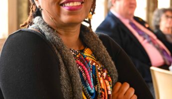 Denise King seated at the speakers table at the Arts Day Luncheon. She is an african american woman with cropped locs. She is smiling, wearing glasses, a black cardigan over a colorful dress with a gray sweater.