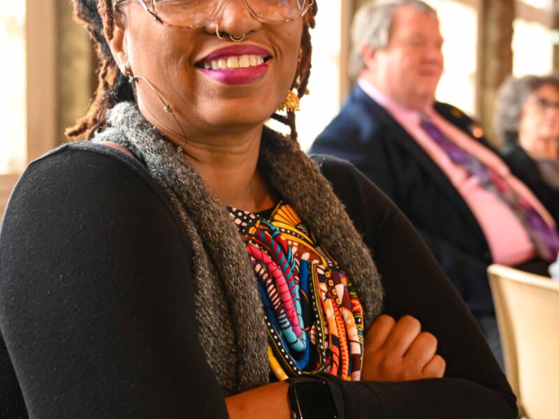 Denise King seated at the speakers table at the Arts Day Luncheon. She is an african american woman with cropped locs. She is smiling, wearing glasses, a black cardigan over a colorful dress with a gray sweater.