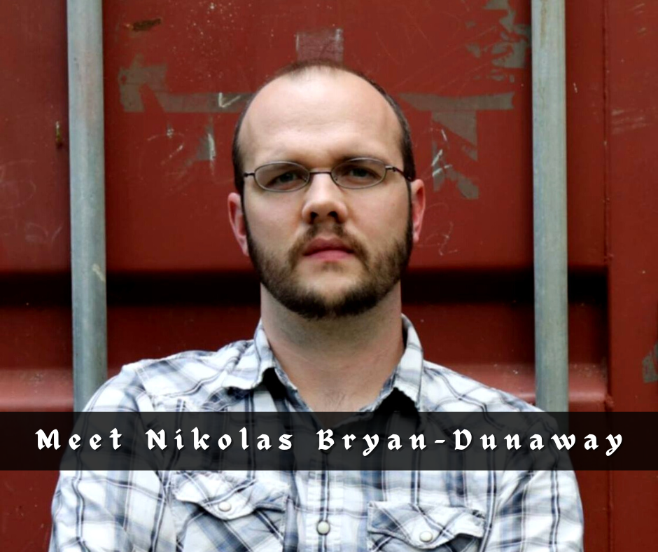 Nikolas Bryan-Dunaway. A Caucasian man with dark hair, thining on top. He has a beard. He is wearing glasses and a plaid button-down shirt. He is standing in front of an industrial storage container that is a rusted red color.