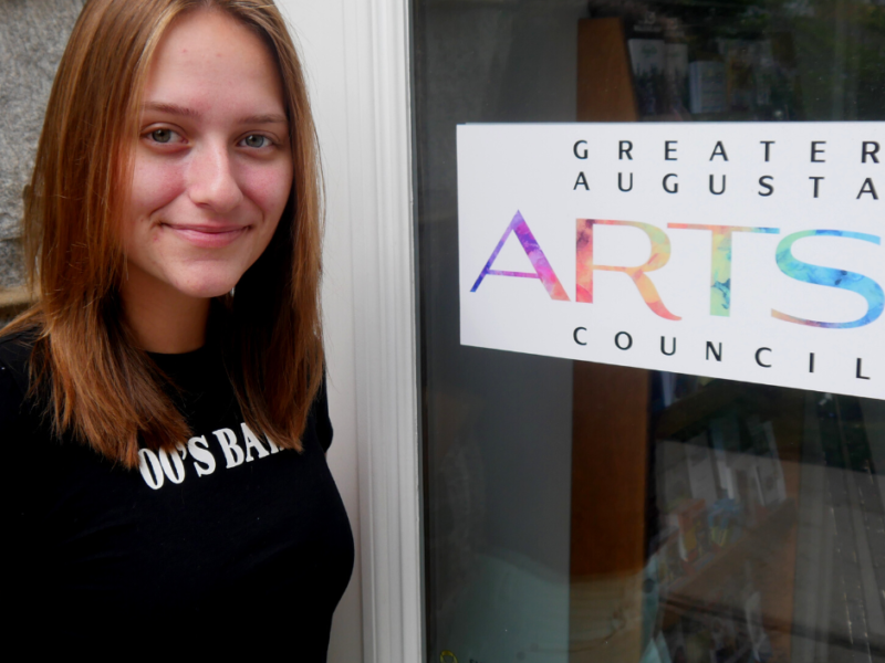 Cameron Thomas standing in front of the greater Augusta Arts council office doors. She is wearing a black tshirt that says "2000s baby" on it. She is caucasian with blue eyes and long, light brownhair. She is smiling.