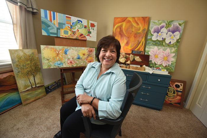 Photo of Ana Thompson sitting in her studio, surrounded by her paintings on easels. Shes wearing a light blue button down shirt with black paints. Her arms are crossed at the wrist. She has short dark hair with bangs. She is smiling.