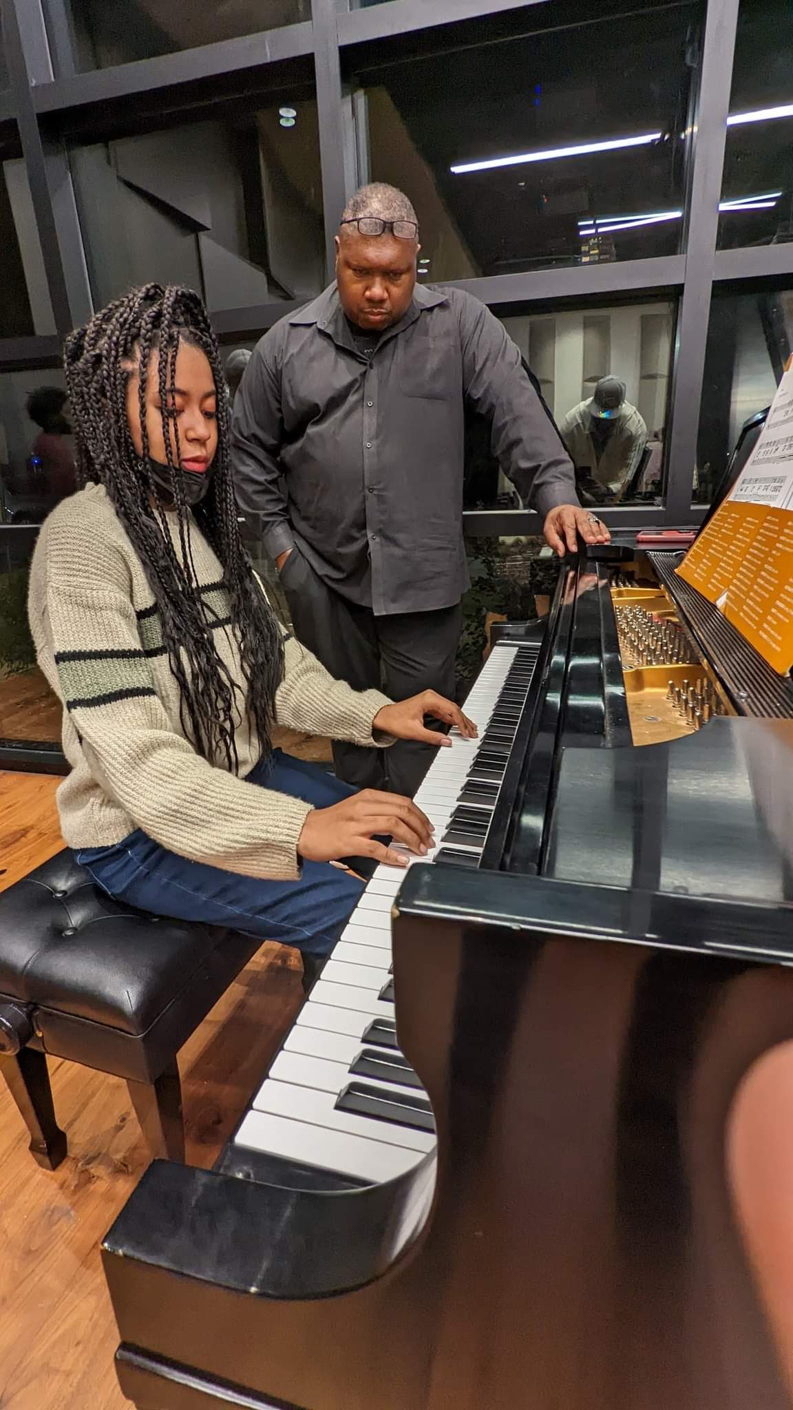 Laurin Mccoy with Wycliff Gordon. She is a young african american teenager with long, black braids. She is seated infront of a piano, that she is playing. She is wearing a beige sweater and blue jeans. Wycliff is standing beside her, teaching her to play. He is an middle aged african american man. He has a shaved head. He is wearing a black button down shirt.
