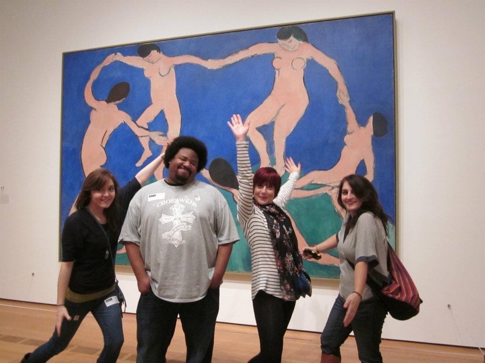 Heather Dunaway, Ernest Anderson, Katie Turner and Emily Smith in front of Henri Matisse's "Dance". On the far left, Heather is a Caucasian woman that has brown hair, swept to the side. She is wearing a navy blue off the shoulder sweater with denim jeans and brown boots. Ernest, an African American man, is taller than Heather. He has a short afro hair style and is wearing a blue and white plaid shirt with baggy denim jeans. His hands are tucked into his pockets and he is laughing. To Ernest's left is Katie. She is a Caucasian woman. she has herhands in the air and she's smiling. She has a short, dyed red pixie cut. She is wearing a white sweater with a rainbow colored scarf and black denim jeans. Emily, who is at the far right, is a Caucasian woman. She is standing awkwardly to be funny. She has long, dark brown hair. She is wearing a gray tshirt, and blue jeans. She has a large brown bag hanging from her shoulder.