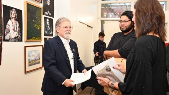 Drake White, an older Caucasian man with a white beard, longer gray hair (slicked back). He is wearing glasses, a navy blue tweed suit jacket and white shirt. He is standing in a gallery featuring his Photographic portraits of Augusta Artists. He is talking to Leslie Hamrick, a Caucasian woman with shoulder length brown hair dressed in black and Devon Lovitt, an African American male with long curly hair and glasses also wearing black.