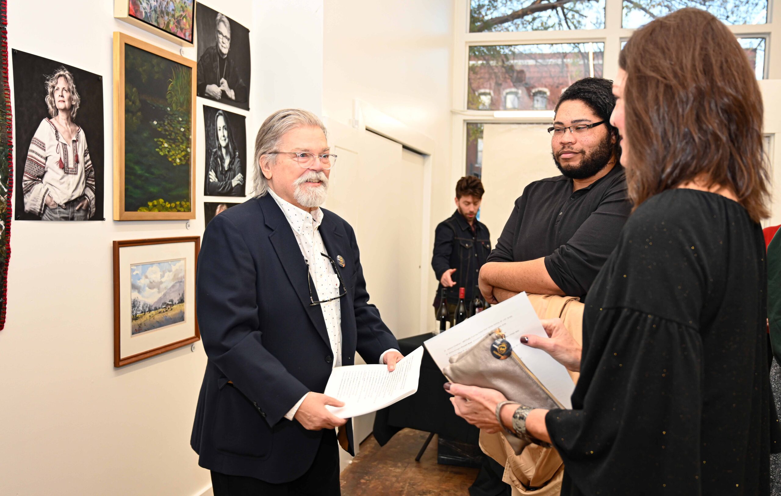 Drake White, an older Caucasian man with a white beard, longer gray hair (slicked back). He is wearing glasses, a navy blue tweed suit jacket and white shirt. He is standing in a gallery featuring his Photographic portraits of Augusta Artists. He is talking to Leslie Hamrick, a Caucasian woman with shoulder length brown hair dressed in black and Devon Lovitt, an African American male with long curly hair and glasses also wearing black.