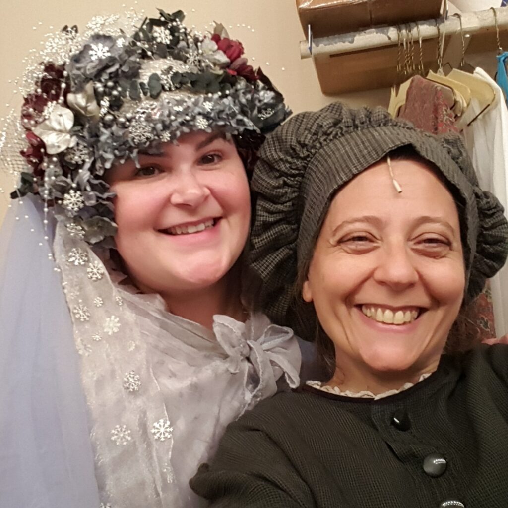 Photo of two women. Katie Reagan on the left. She is dressed as the Ghost of Christmas Past from the Christmas Carol. SHe has dark hair and is wearing a snowy floral crown and white lacey dress. Pax Bobrow, on the right, is dressed as Mrs. Mops from A Christmas Carol wearing a floppy brown hat and dark dress with a white collar. Both women are smiling.
