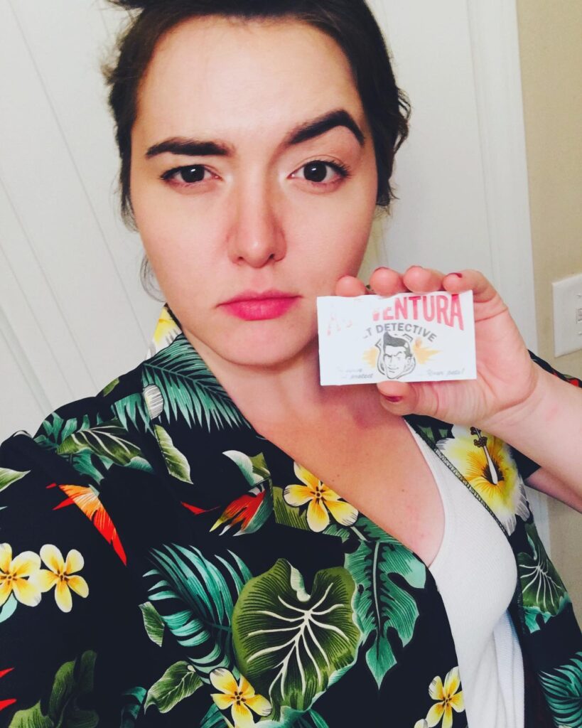 Heather Dunaway dressed as Ace Ventura, Pet Detective for Halloween. She is wearing a Hawaiian shirt, white tank top and black and red striped pants.