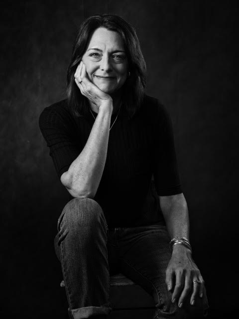 Photo of Leslie Hamrick taken by Drake White. A black and white image with heavy contrast. Leslie is wearing a dark sweater. She is a Caucasian woman with shoulder length brown hair. She is half sitting on a stool facing the camera. She has her right hand tucked under her chin and propped up on her right knee. Her left hand is on her left thigh. She is smiling.