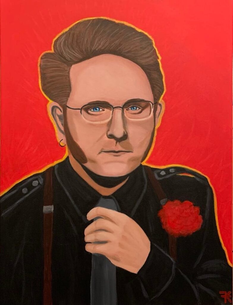 Portrait of Paul Marsh by Francie Klopotic. He is infront of a red backdrop wearing a black suit with a red flower. He is adjusting his tie. Paul is a Caucasian man with brown hair and sideburns. He is wearing glasses.