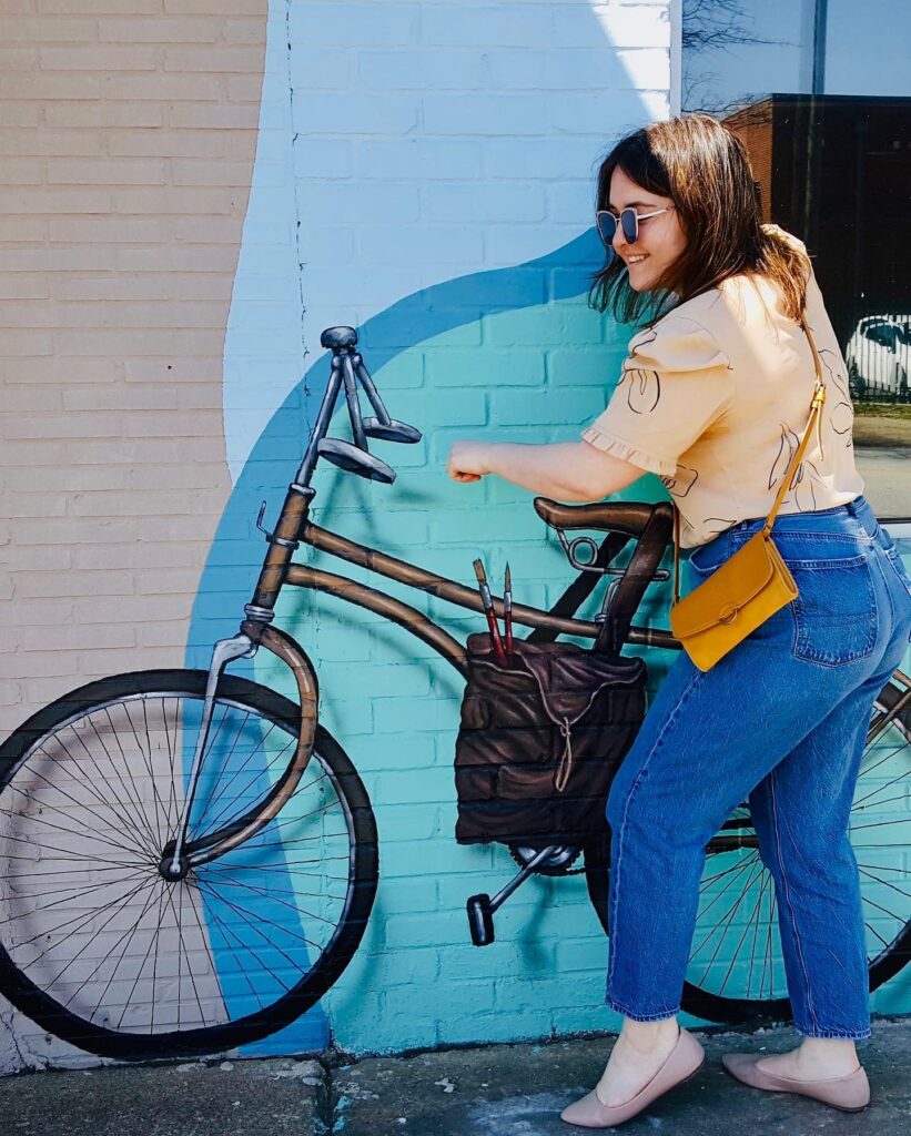 Heather Dunaway pretending to mount a painting of a bicycle. Heather is a Caucasian woman with shoulder length brown hair. She is wearing sunglassese, a beige short sleved blouse and blue jeans.