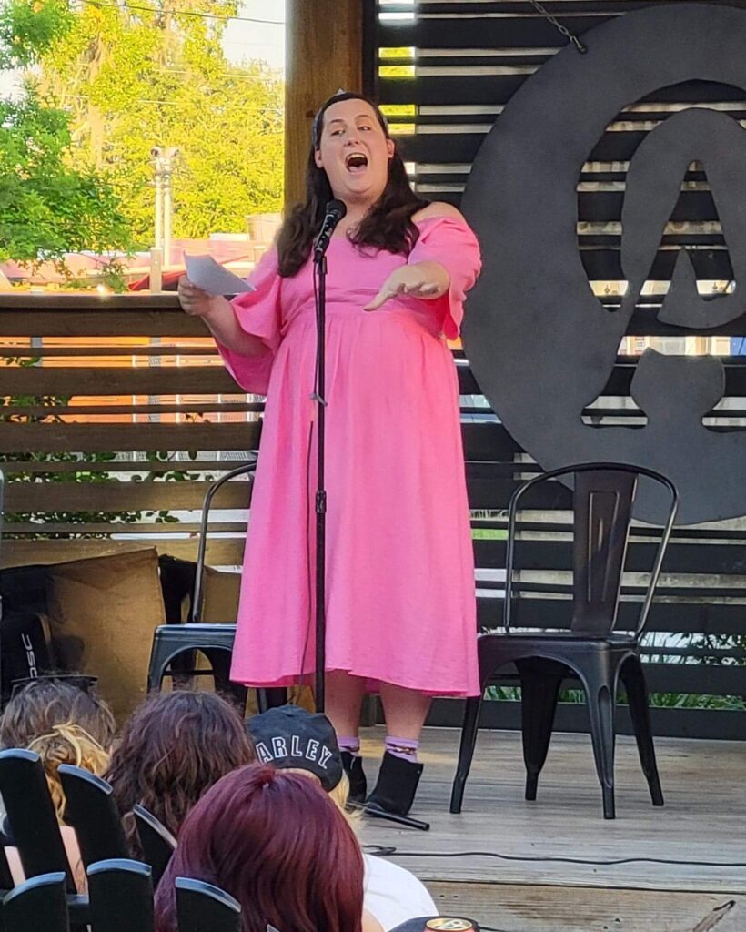Evelyn Berry, giving a spoken word performance. She is a Caucasian woman who has long dark hair. She is wearing a pink floral dress and black boots.