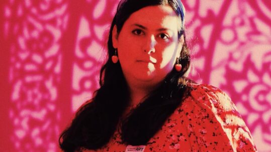 Evelyn Berry is a Caucasian woman with long dark hair and dark eyes. She is wearing pink earrings and a pink floral dress. The backdrop is pink flowers.