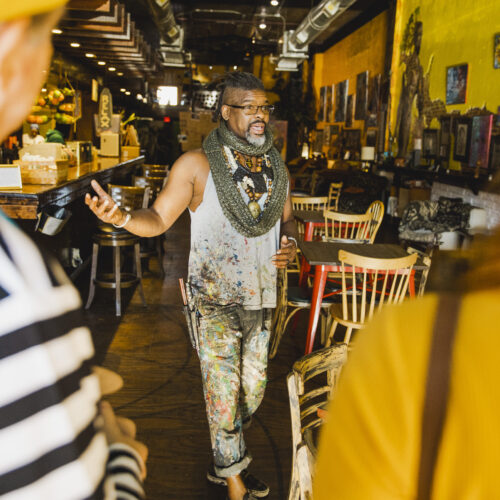 Baruti Tucker, an African American Male wearing paint splattered jeans, tank top and scarf. His hair is dreaded and pulled back into a half-up-do. He is wearing glasses standing in Humanitree House, his juice bar and restaraunt. The walls are yellow and covered in local art.