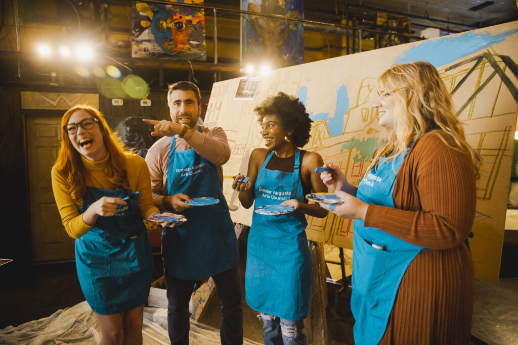 Photo of a group of friends painting and laughing. All are wearing bright blue aprons and holding brushes with paint pallets. From left to right we see a red-headed Caucasian woman wearing a yellow sweater, an older Caucasian man with salt-and-pepper hair wearing a brown shirt, an African-American woman with natural hair and a black tank top, and a blonde Caucasian woman wearing a light brown dress.
