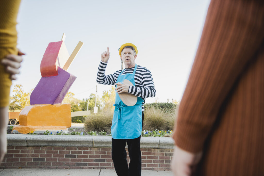 A Caucasian man with a yellow barrette, black and white striped shirt and bright blue painters apron standing in front of a popsicle sculpture, pointing up.
