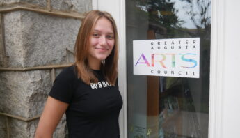 Cameron Thomas is a Caucasioan woman with shoulder length light brown hair and blue eyes. She is wearing a black tshirt standing in front of the Arts Council door with the Greater Augusta Arts Council Logo.