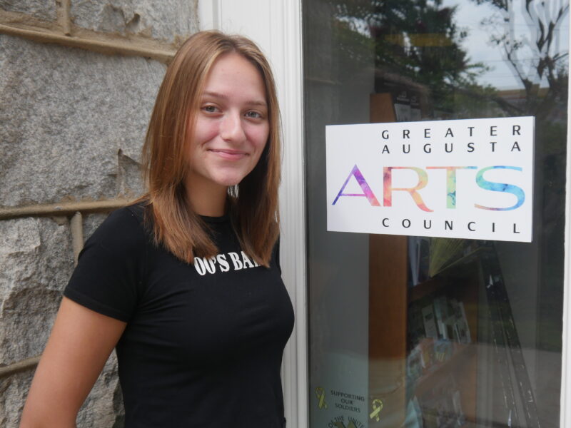 Cameron Thomas is a Caucasioan woman with shoulder length light brown hair and blue eyes. She is wearing a black tshirt standing in front of the Arts Council door with the Greater Augusta Arts Council Logo.