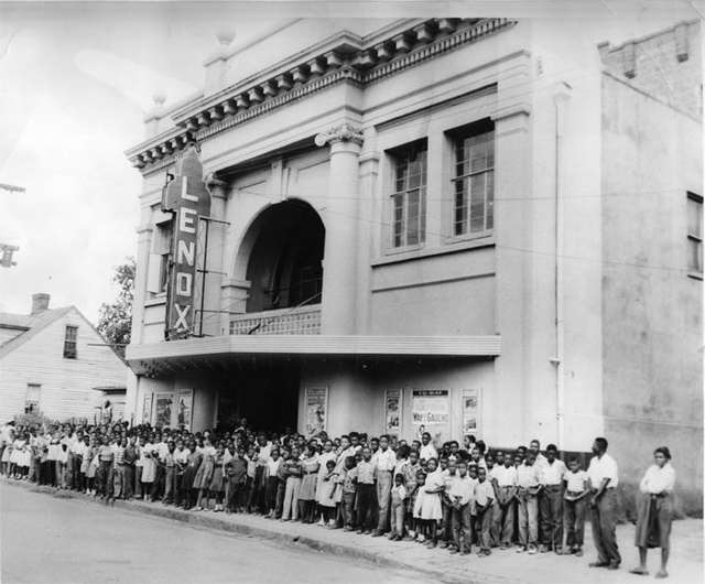 Photo of the Lenox Theatre. A marble two story building with large windows and LENOX in boxed lettering down the side. It has a line of people out the door, waiting to enter for a show.