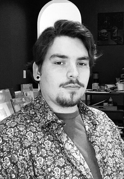 A black-and-white photo of Logan Beasley. He is a Caucasian man with dark hair, gauged ears and a mustache/goatee. He is wearing a floral printed button down with a shirt underneath.