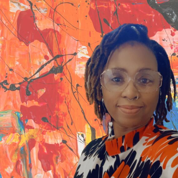 Photo of African American woman standing infront of a painting. She is wearing a colorful dress, earrings and glasses.