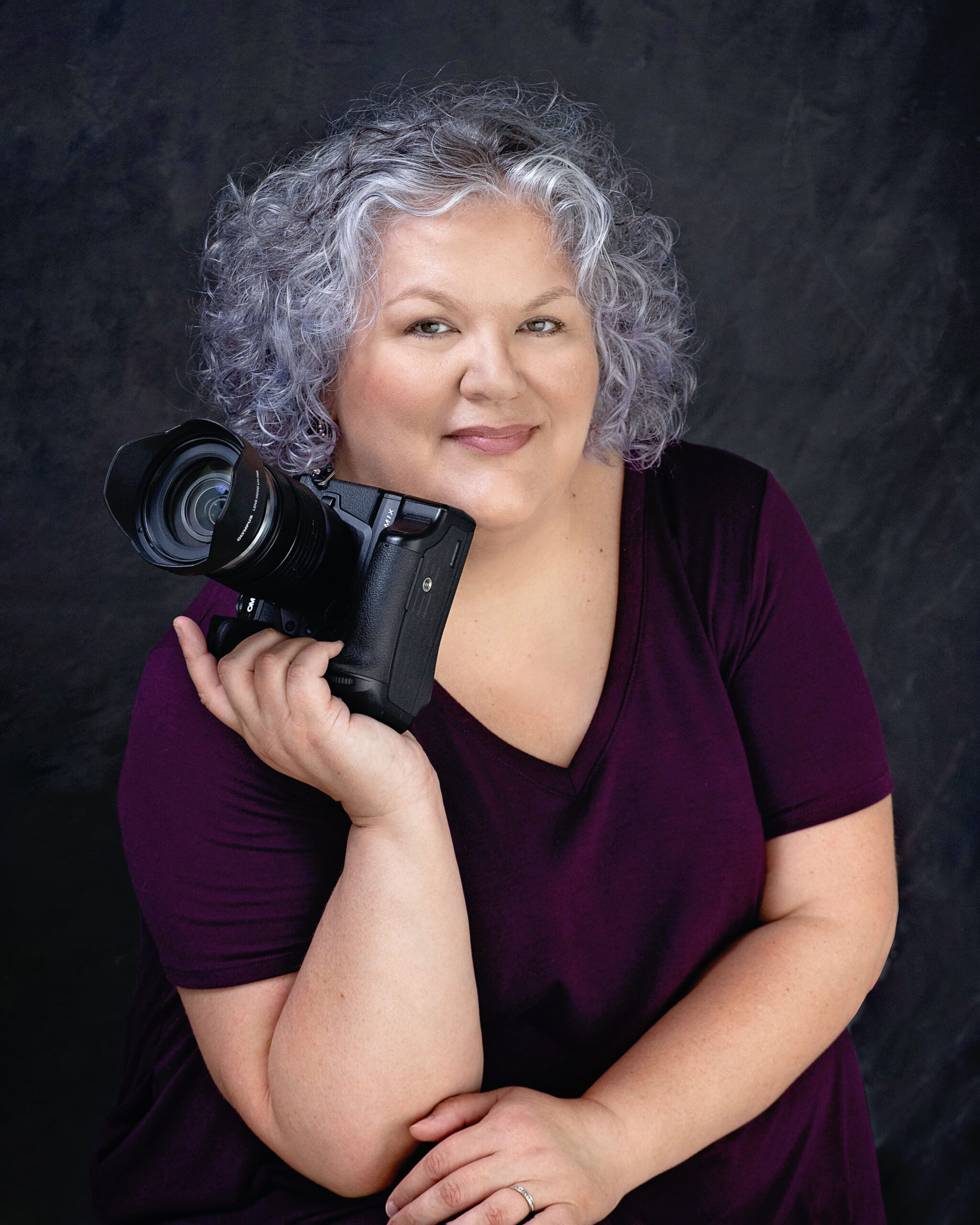Cassandra Bayer portrait. A woman with gray/white hair in a black tshirt holding a DSLR camera.