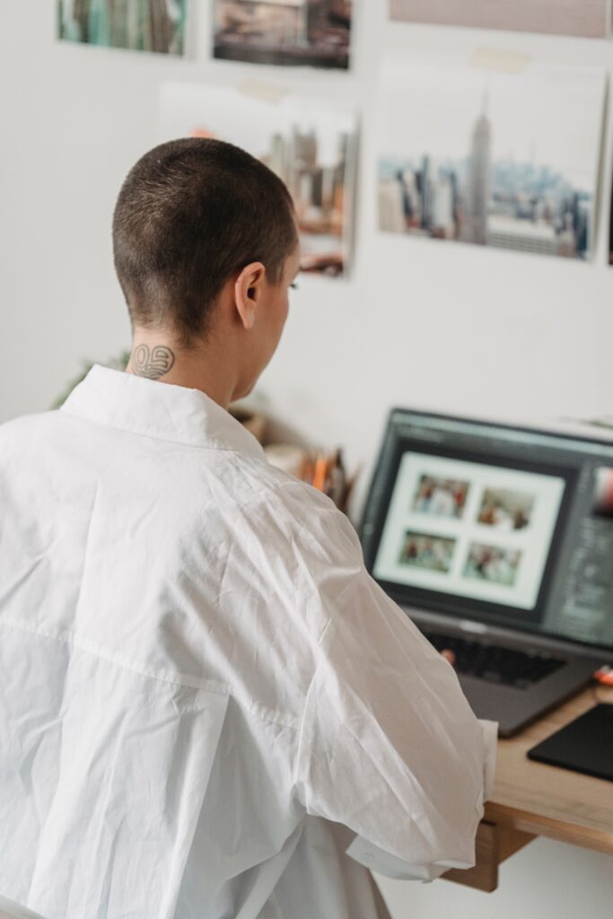 Photo of a girl with a shaved head wearing a large white shirt. She has her back to the camera as she scrolls on her computer in her art studio.