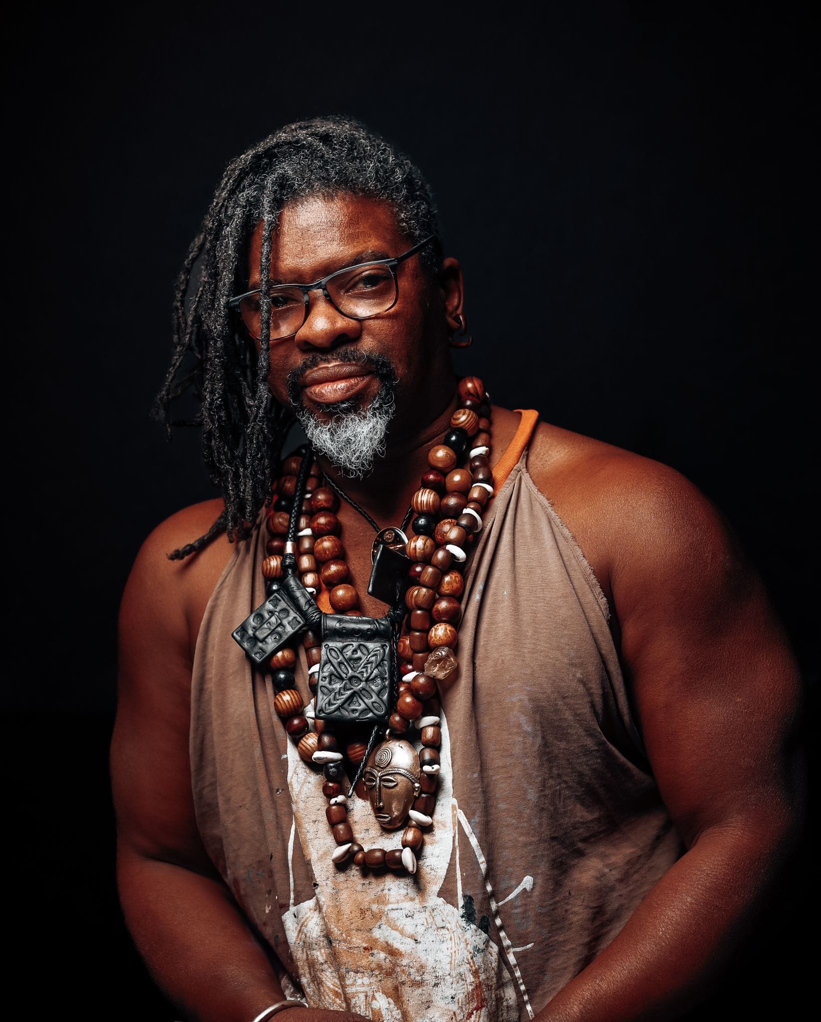 Photo of Baruti. He is an African American man with shoulder length dreads and a graying goatee. He is wearing a paint stained brown tank top and bead necklaces.