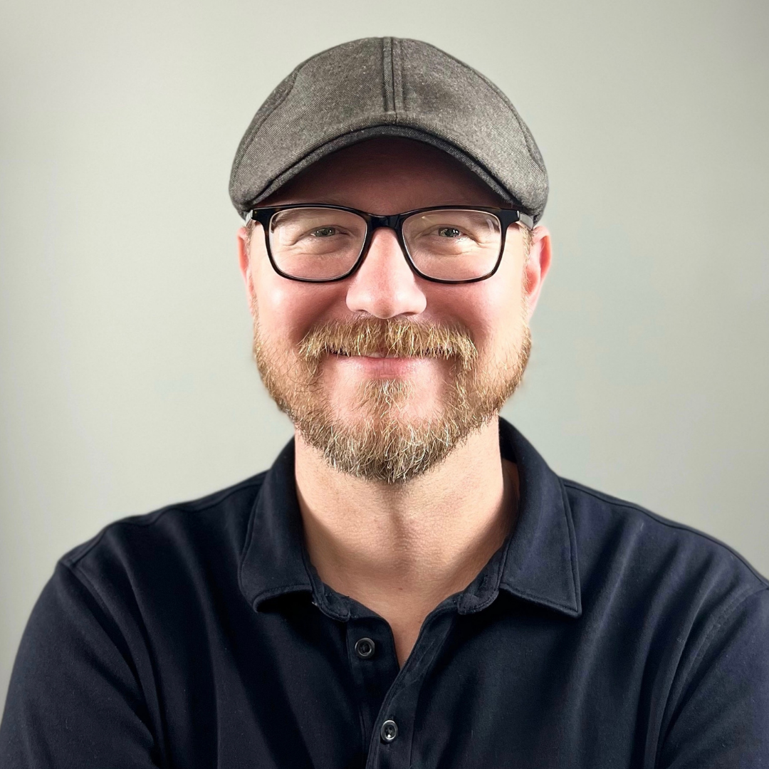 Portrait of Jason. He is a Caucasian man with light colored beard, black glasses. He is wearing a grey hat and black collared shirt.