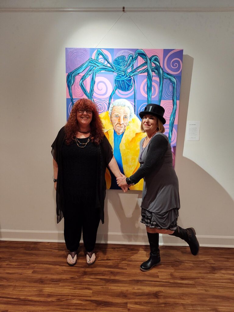 Photo of Brenda Durant and Rhian Swain standing in front of Rhian's artwork at her show "Breaking the Barriers". Rhian is a Caucasian woman with red curly hair, she is wearing all black. Brenda is a Caucasian woman with short blonde hair, she is waring a black hat and gray dress with black cardigan. Both women are smiling.