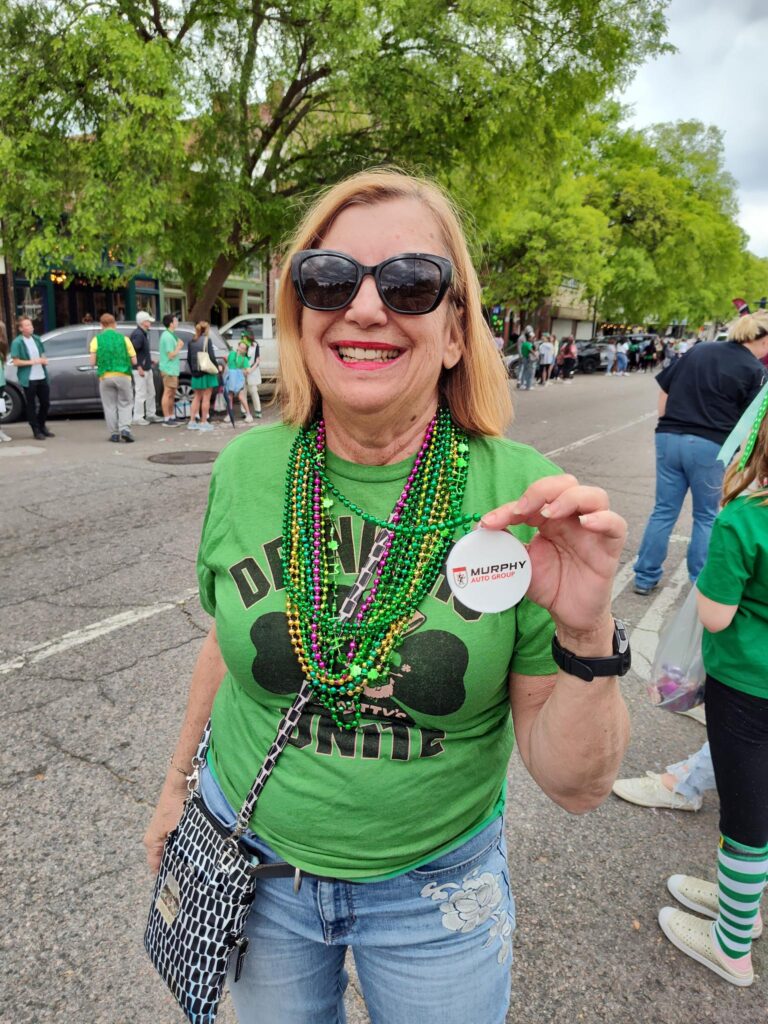 Brenda Durant, dressed ina green tshirt with a four leaf clover printed on it. She is wearing green festival beads. She is a Caucasian woman with blond hair. She is smiling, wearing sunglasses.