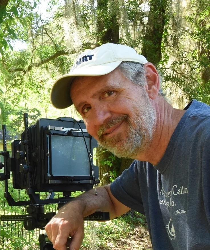 Photo of Mark. He is a Caucasian man with gray hair and beard. He is wearing a white baseball cap and blue collared shirt. He is standing outside with his camera on a tripod.