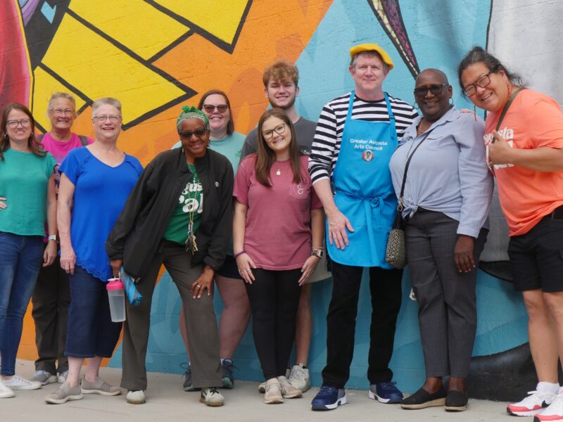 Photo of Promenade & Paint tour group. There are 8 women and two men. All of different backgrounds wearing different vibrant colors. They are standing in front of a colorful James Brown Mural smiling.