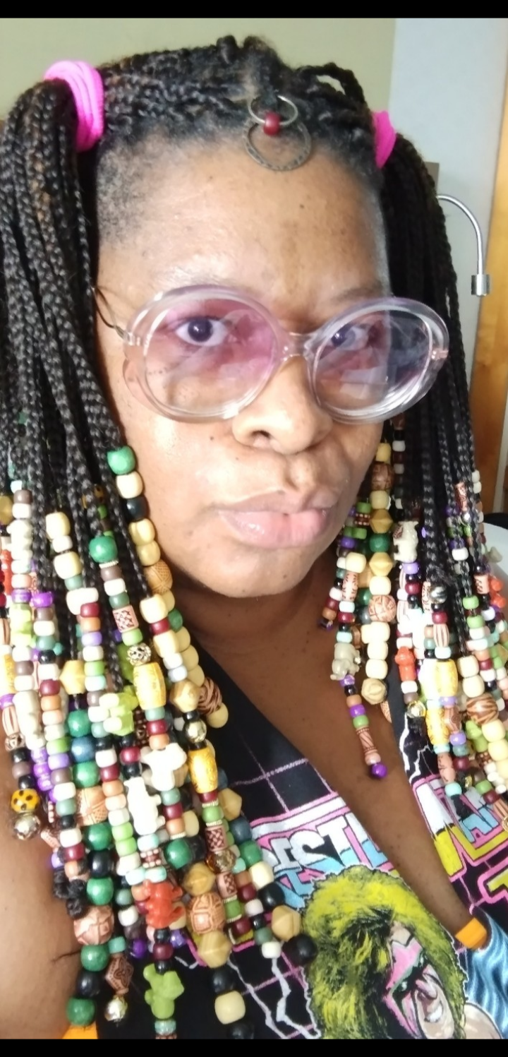 Photo of Judi. She is an African American woman with dark, dreaded hair cropped at her shoulders. She is wearing pink glasses and a floral print top.