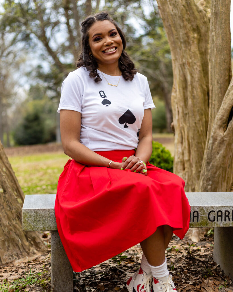 Crystal Jones portrait. She is a woman with dark brown curly hair. She is sitting on a bench. She is wearing a red skirt and white, queen of spades tshirt. She is smiling with her hands clasped in her lap.