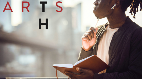 Photo of an African American man with dreads in a poly tail. He is wearing ear pods, a white tshirt layred with a black button down. He is writing in a leather bound book while looking over a city scape.
