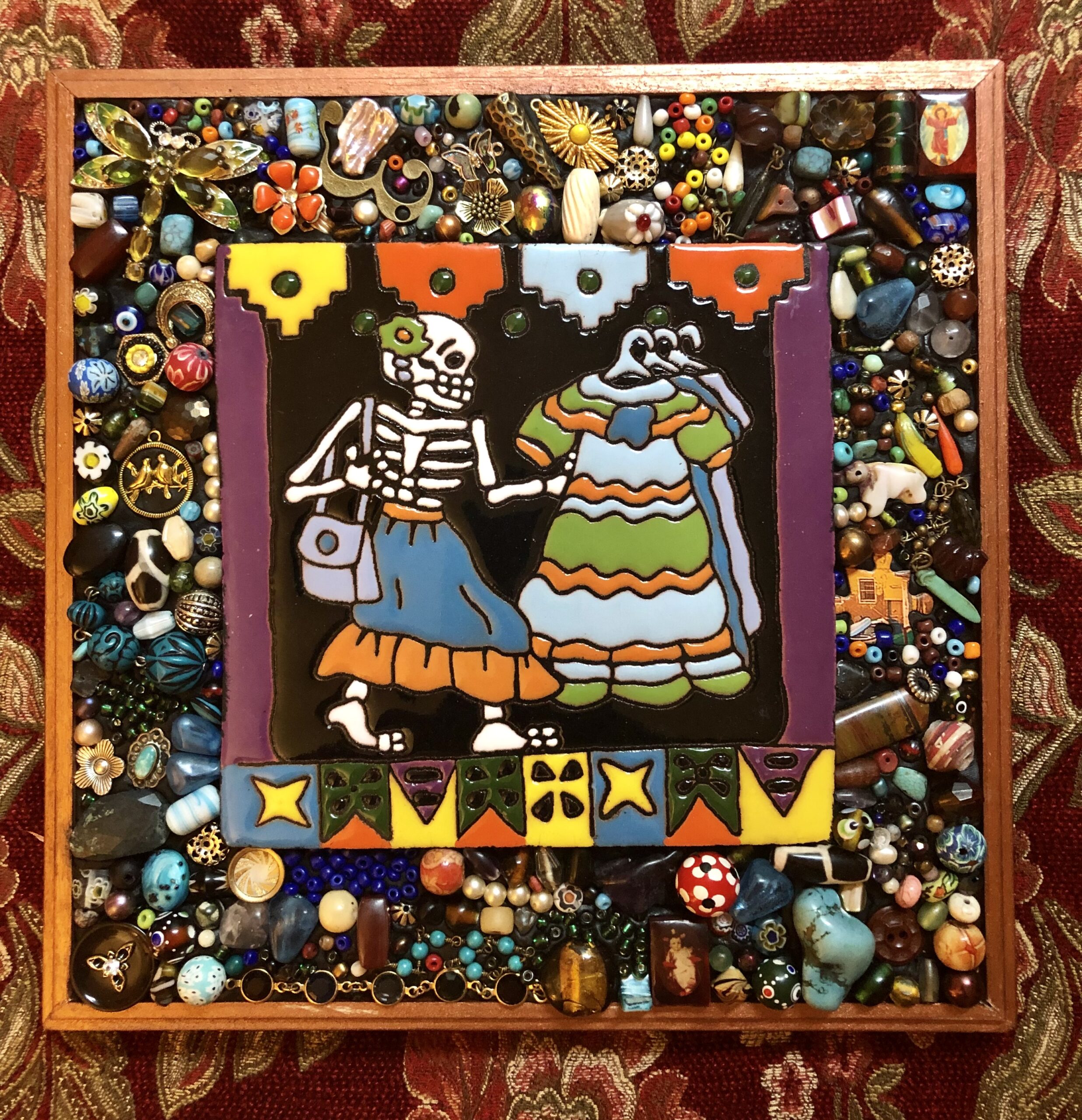 The centerpiece of this artwork by Laurie Algar is a mosaic of a skeleton in a skirt looking at a rack of dresses, surrounded by a frame filled with small pieces of colorful jewelry and beads.