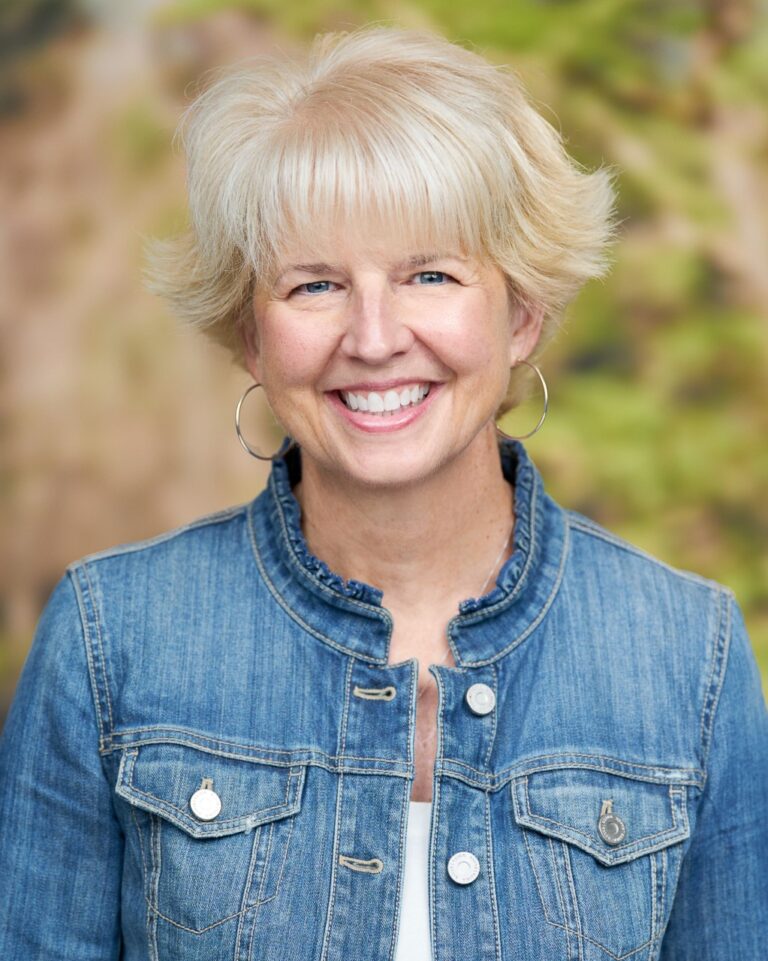 June is a Caucasian woman with short, white blonde hair. She is wearing a blue denim jacket and white shirt. She is smiling.
