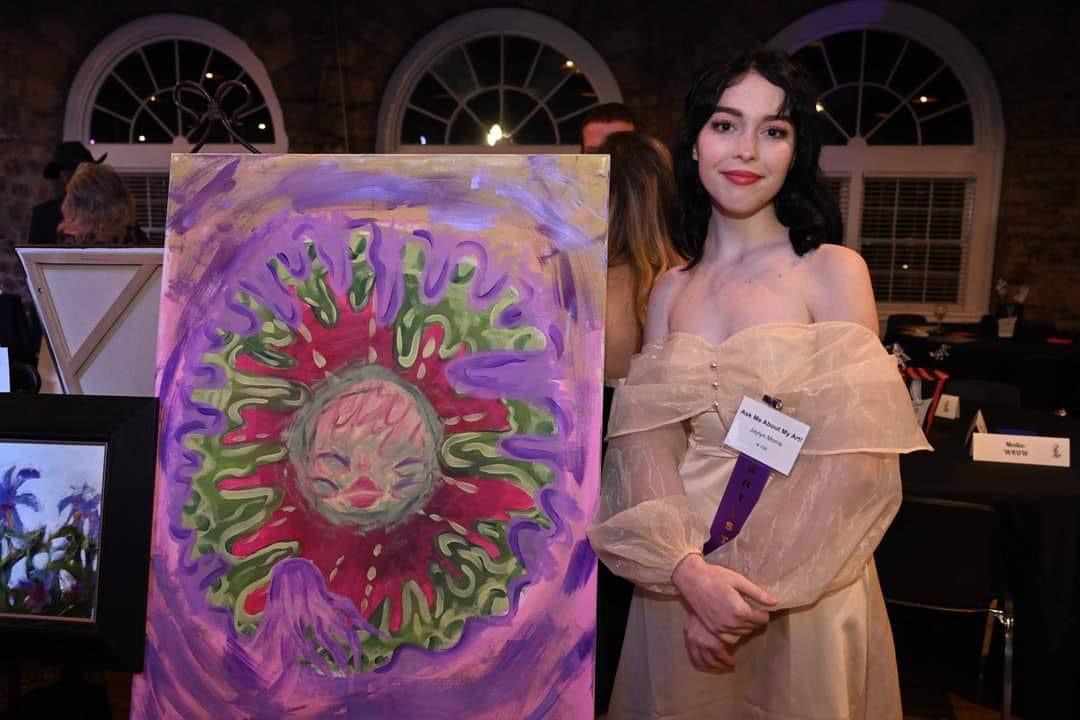 Photo of a Caucasian woman standing next to a purple painting. She has black hair and is wearing a beige poofy dress