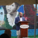 Corey Rogers giving a speech. He is an expressive African-American man who is bearded, bald, and is wearing a dark blue suit