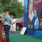 Pax Bobrow, Greater Augusta Arts Council Project Manager, giving a speech. She is a short Caucasian woman with long brown hair and wearing an aqua-colored top and dark blue pants