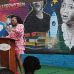 Salonika Rhyne, mural artist, giving a speech. She is an African-American woman with a bright pink dress and poofy hair.