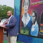 Corey Rogers giving a speech. He is an expressive African-American man who is bearded, bald, and is wearing a dark blue suit