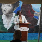 Judge Monique Walker giving a speech. She is an African-American woman with short hair, matching white suit and pants, and a yellow top