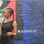 Nefertiti Robinson singing in front of Golden Blocks mural. She is an African-American woman in a black dress with braided hair
