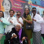 Group photo including Ruth B. Crawford in front of the mural. All women are smiling and holding up their pinky